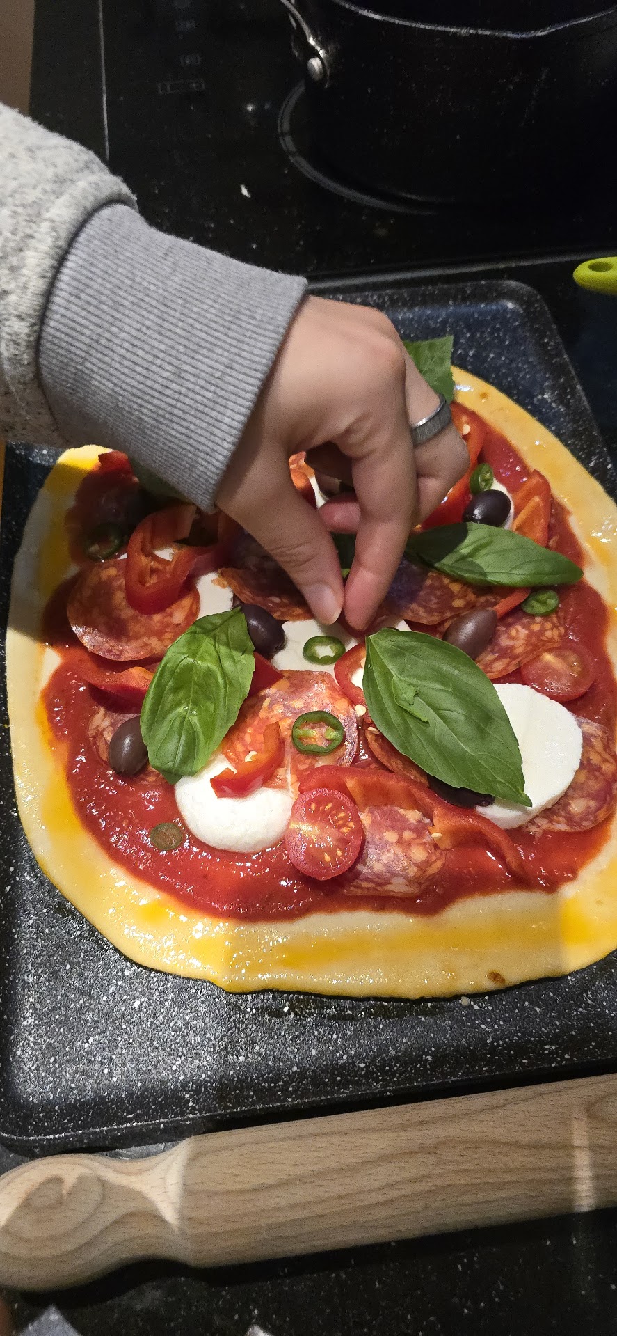 Hand placing fresh basil onto a homemade pizza topped with pepperoni, mozzarella, olives and chillies
